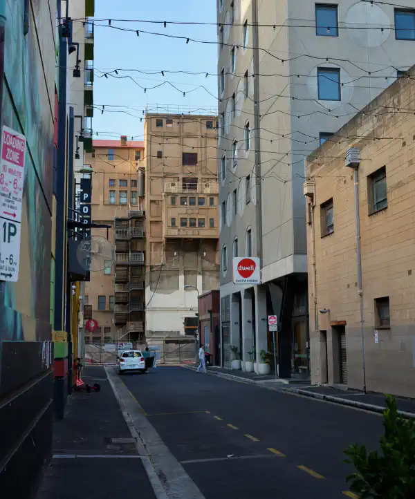 View down a short city side street. Lights are strung across it. There are buildings surrounding it on all sides.
